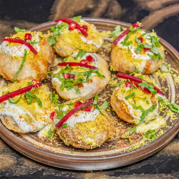 Plate of round, fried Indian snacks topped with creamy sauce, crunchy yellow bits, green herbs, and red garnishes.