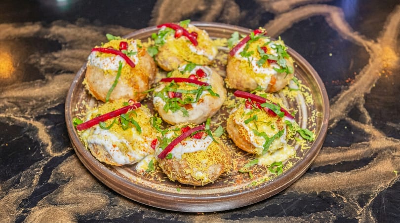 Plate of round, fried Indian snacks topped with creamy sauce, crunchy yellow bits, green herbs, and red garnishes.