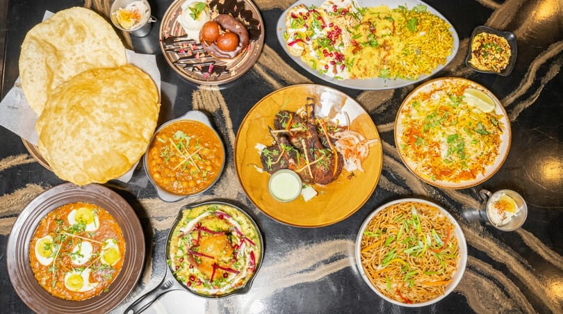 A spread of vibrant Indian cuisine, including chickpea curry, puri, lamb chops, biryani, and desserts.