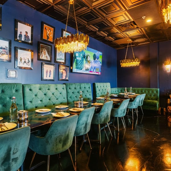 Dark-walled restaurant with green velvet seating, framed art, set tables, and sparkling gold chandeliers.