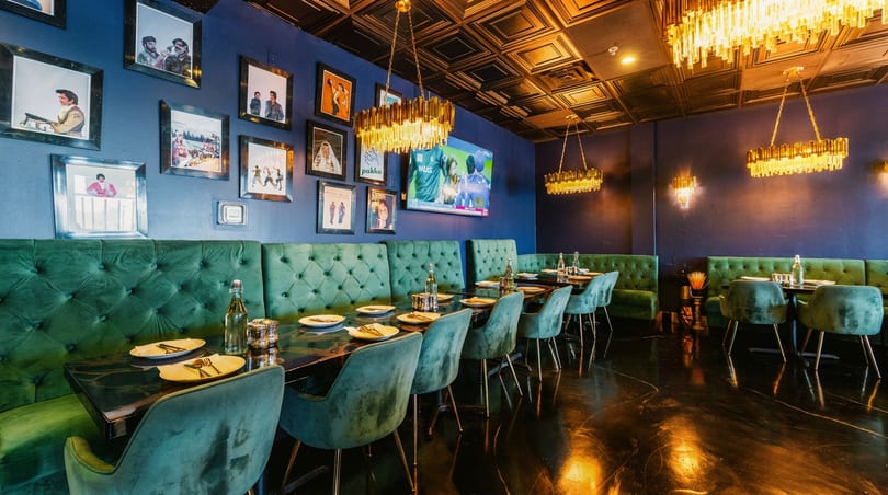 Elegant restaurant interior with dark blue walls, green velvet seating, gold chandeliers, and framed art.
