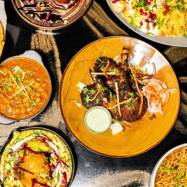 Overhead view of a vibrant spread of Indian dishes: curries, rice, fried bread, lamb chops, and noodles.