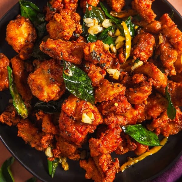 Crispy fried chicken pieces on a black plate, garnished with curry leaves, garlic, and chilies.