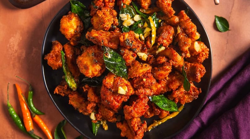 Spicy fried chicken pieces with green chilies, curry leaves, and garlic on a black plate.