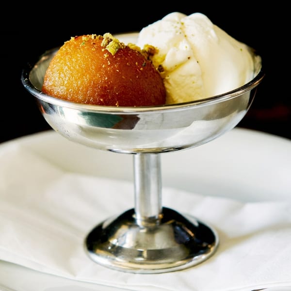 Gulab jamun with pistachios and vanilla ice cream in a silver bowl on a white plate.