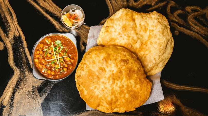 Chana masala with two fried flatbreads, sliced onions, and lemon on a dark, patterned table.