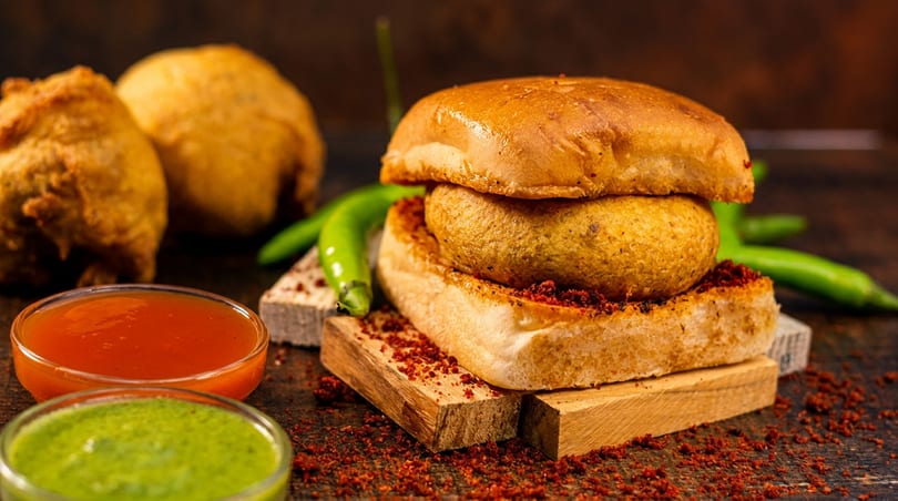 Vada Pav with green chilies, red spice, and two bowls of chutney on a rustic wooden surface.