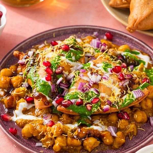 Colorful Indian chaat with chickpeas, sauces, pomegranate, and crispy toppings on a purple plate.