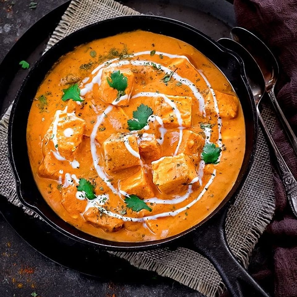 Rich orange paneer curry with cream and cilantro in a cast iron pan, with spoons and cashews.