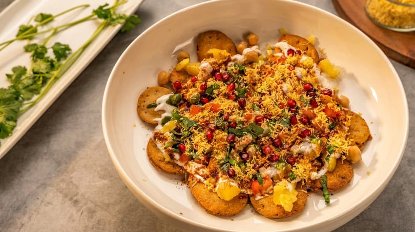 Colorful Indian papdi chaat with yogurt, chutneys, pomegranate, and sev in a white bowl.