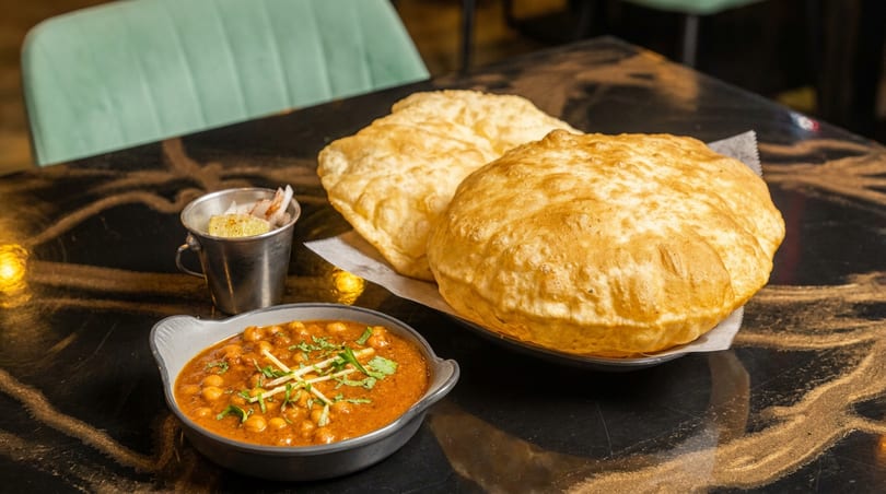 A bowl of chana masala with cilantro, two large bhature, and a side of lemon.