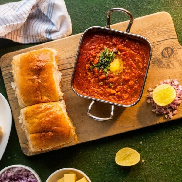 Pav bhaji with butter and cilantro, served with golden bread rolls, chopped onions, and lemon.