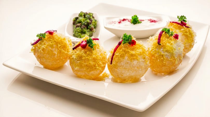 Five golden pani puri topped with sev, pomegranate, and herbs, served with green and white dips.