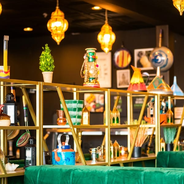 Vibrant restaurant interior featuring golden shelves with colorful decor, ornate lamps, and green velvet seating.