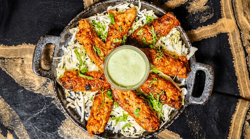 Grilled chicken kebabs on shredded cabbage with a green dipping sauce in a rustic pan.