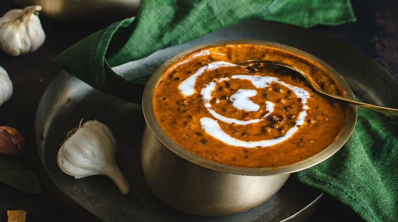 Bowl of creamy dal makhani with a golden spoon, garlic cloves, and a green napkin on a dark tray.
